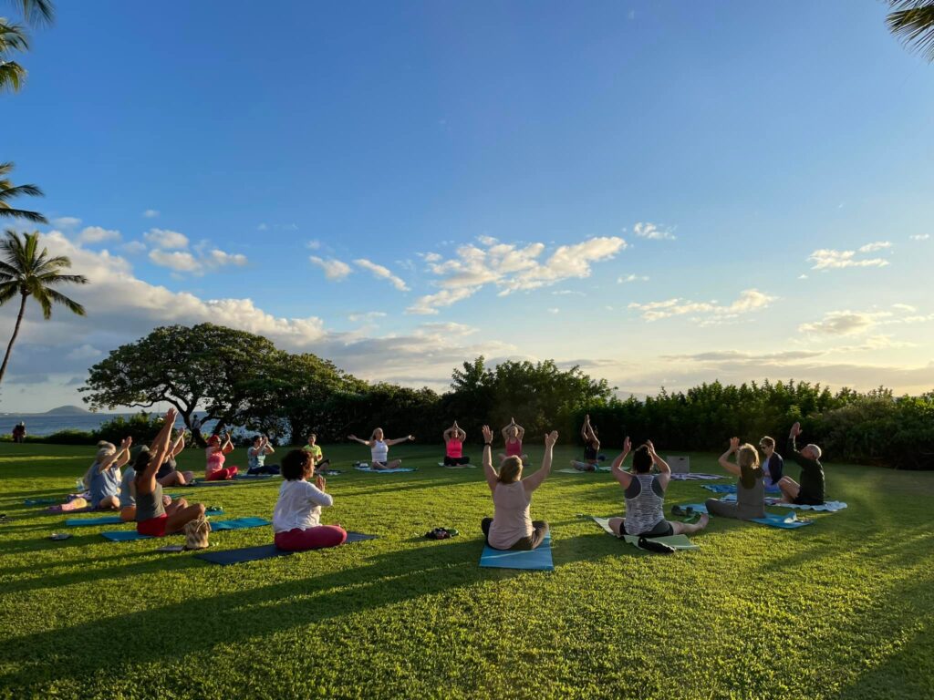 Tanya Proffitt leading a serene outdoor group meditation on a lush lawn overlooking the ocean at sunset during a yoga retreat.