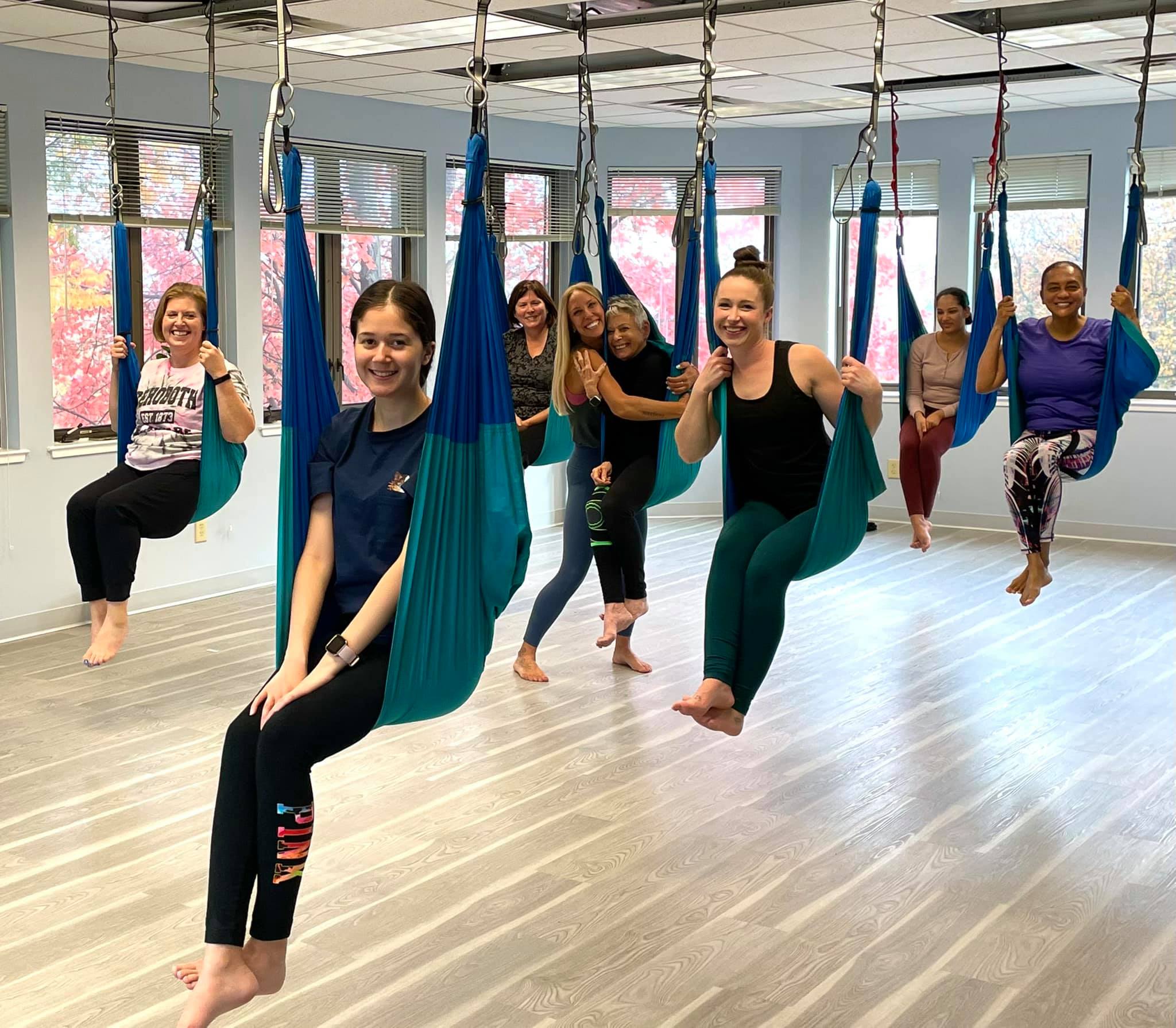 Tanya Proffitt with a group of smiling women practicing aerial yoga in blue hammocks at a sunny studio in Olney, Maryland.